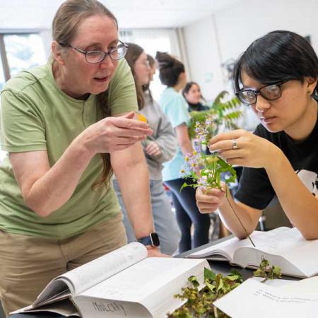 A woman professor leans over a lab table and gestures to a purple-flowered plant that a student is holding. There are several open books on the table to help the two identify the plant.